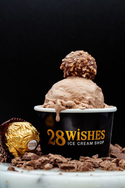 A scoop of chocolate-brown ice cream topped with a nut-covered chocolate truffle in a black 28 Wishes cup, with a gold-wrapped chocolate and chocolate pieces beside it